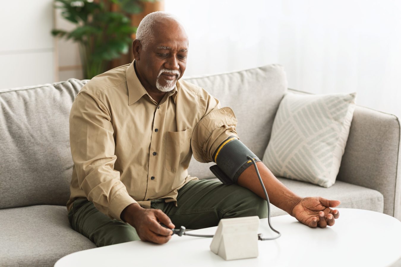 Mature African American Man Measuring Arterial Blood Pressure With Sphygmomanometer Medical Device Sitting On Couch Indoors. High Blood-Pressure, Hypertension Health Problem Concept